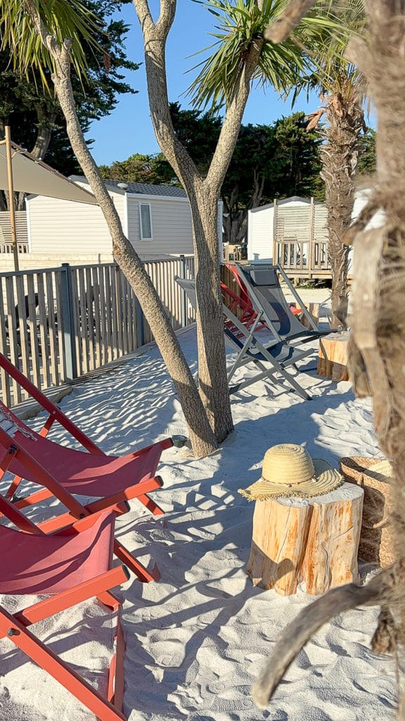 Transats rouges sur plage sable, mobile-homes en arrière-plan.